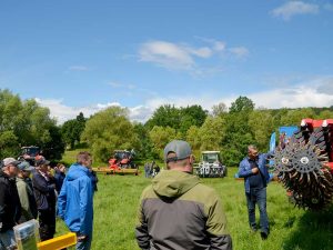 Mehrere Menschen stehen auf einer grünen Wiese vor einer landwirtschaftlichen Maschine. Ein Referent erläutert die Vorführung mit einem Mikrofon.