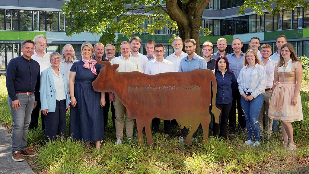Personengruppe steht auf einer Grünfläche; im Vordergrund eine Kuhfigur aus Metall; im Hintergrund ein Baum und ein Gebäude