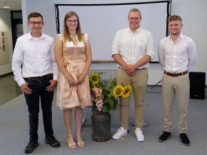 4 Personen stehen in einem Raum; in der Mitte ein großer Blumenstauß