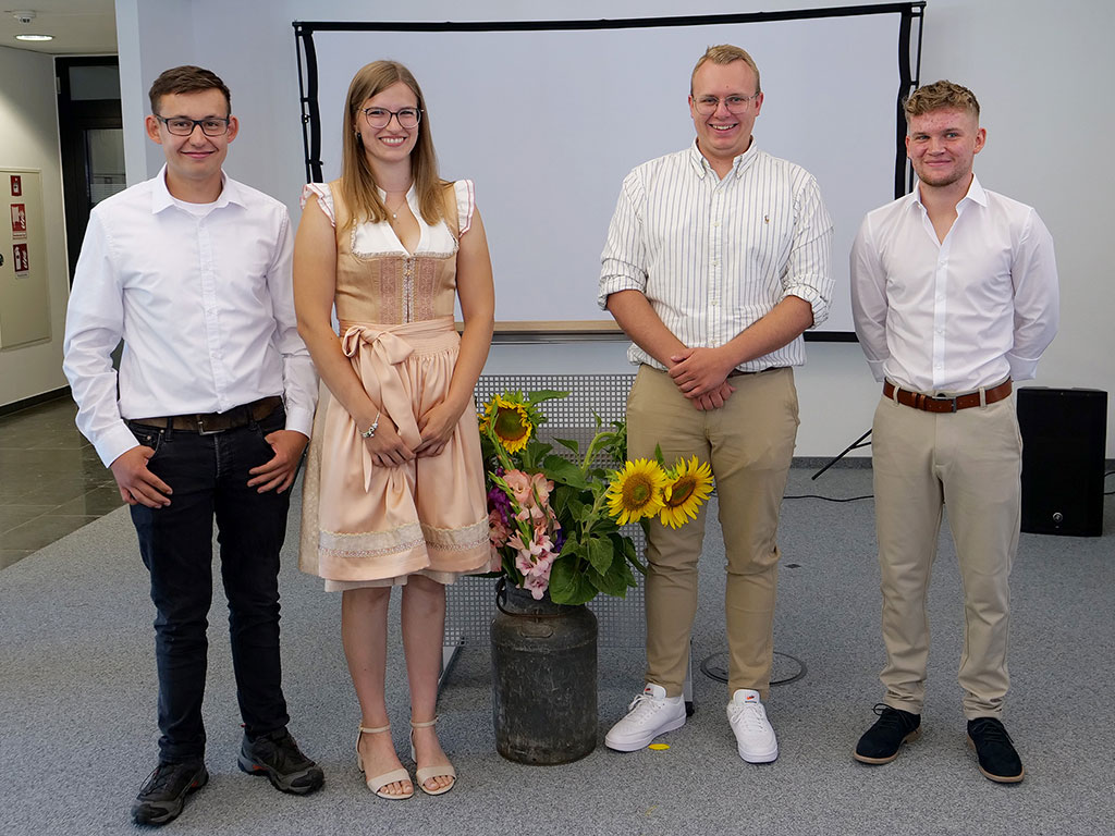 4 Personen stehen in einem Raum; in der Mitte ein großer Blumenstauß