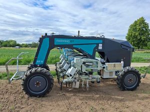 Zu sehen ist ein selbstfahrender Unkrautroboter Farming GT 38 von Agravis. In der Mitte des Geräts sind die Hackgeräte zur mechanischen Unkrautbekämpfung angebracht. Das Gerät steht auf einer Ackerfläche. Im Hintergrund sind ein Getreidefeld, Büsche und Bäume zu sehen.
