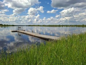Eine Landschaftsaufnahme zeigt einen See, dessen Enden gerade noch zu erkennen sind. Der Betrachter steht am Seeufer, das etwa 2 m über dem Wasserspiegel liegt und von hohem Gras bewachsen ist. In den See ragt ein T-förmiger Steg mit einem Rettungsring. In weiter Ferne hinter dem See ist die Landschaft geprägt von grünen Bäumen und ein paar Windrädern. Der blaue Himmel ist übersät von Schönwetterwolken und spiegelt sich auf der Wasseroberfläche.