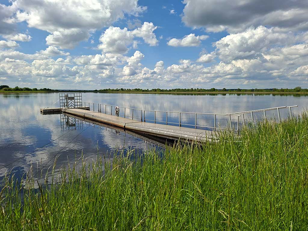 Eine Landschaftsaufnahme zeigt einen See, dessen Enden gerade noch zu erkennen sind. Der Betrachter steht am Seeufer, das etwa 2 m über dem Wasserspiegel liegt und von hohem Gras bewachsen ist. In den See ragt ein T-förmiger Steg mit einem Rettungsring. In weiter Ferne hinter dem See ist die Landschaft geprägt von grünen Bäumen und ein paar Windrädern. Der blaue Himmel ist übersät von Schönwetterwolken und spiegelt sich auf der Wasseroberfläche.
