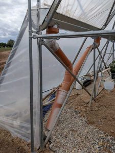 Die Abbildung zeigt ein Y-förmiges Fallrohr aus orangebraunen KG-Rohren. Zwei Enden sind an die Regenrinne angeschlossen. Ein Ende verschwindet in einem Kiesbett auf dem Boden, Hinter dem Rohr und der Rinne ist gespannte Folie und ein Teil der Stahlkonstruktion des Tunnels zu sehen.