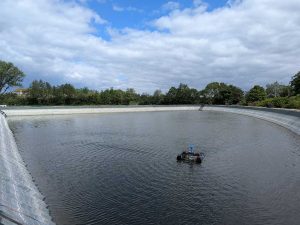 Das Foto ist von der Böschung zu einem großen Teich aufgenommen worden. Bis zur Wasseroberfläche ist die steile Böschung ca. 2 Meter lang und sie ist mit Folie ausgelegt. Der Teich nimmt fast das gesamte Bild ein. Auf dem Teich schwimmt ein Oloid, er sieht aus wie ein Mini-Katamaran und sorgt für die Belüftung des Wassers. Im Hintergrund ist die Szenerie von Bäumen eingerahmt. Am Himmel sind weiße Wolken zu sehen, sie geben in einem kleinen Ausschnitt den Blick auf den blauen Himmel frei.