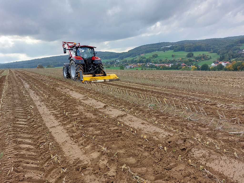 Traktor auf Maisstoppelfeld, im Hintergrund hügelige Landschaft mit Wohnbebauung und Wald