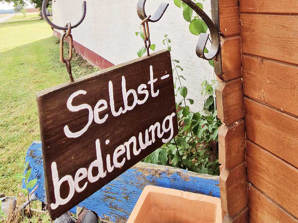 Das Bild zeigt ein Schild vor einer Gartenlaube mit der Aufschrift Selbstbedienung.
