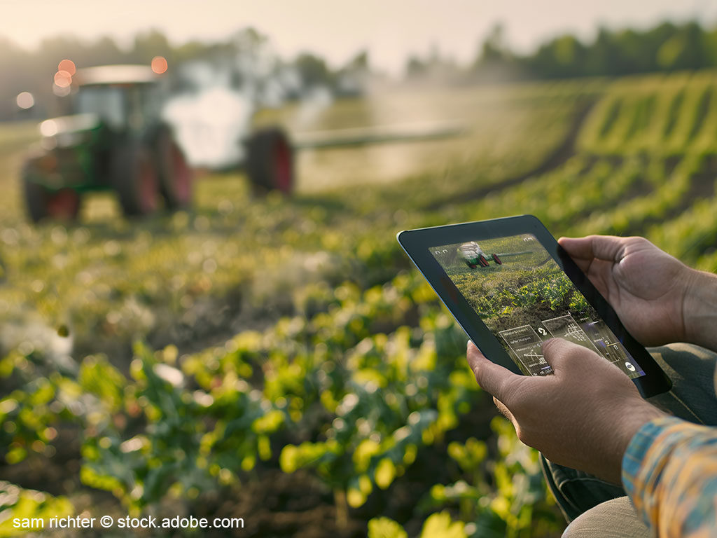 Man die Hände eines Landwirtes,welche ein Tablet halten; im Hintergrund ein Trecker bei der Feldarbeit