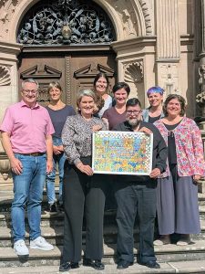 Auf einer Treppe steht eine Personengruppe in 2 Reihen. Die beiden Personen in der vorderen Reihe halten gemeinsam eine große bunte Urkunde in den Händen. Im Hintergrund sieht man eine schöne verzierte Eingangstür aus dunklem Holz eines alten Gebäudes.