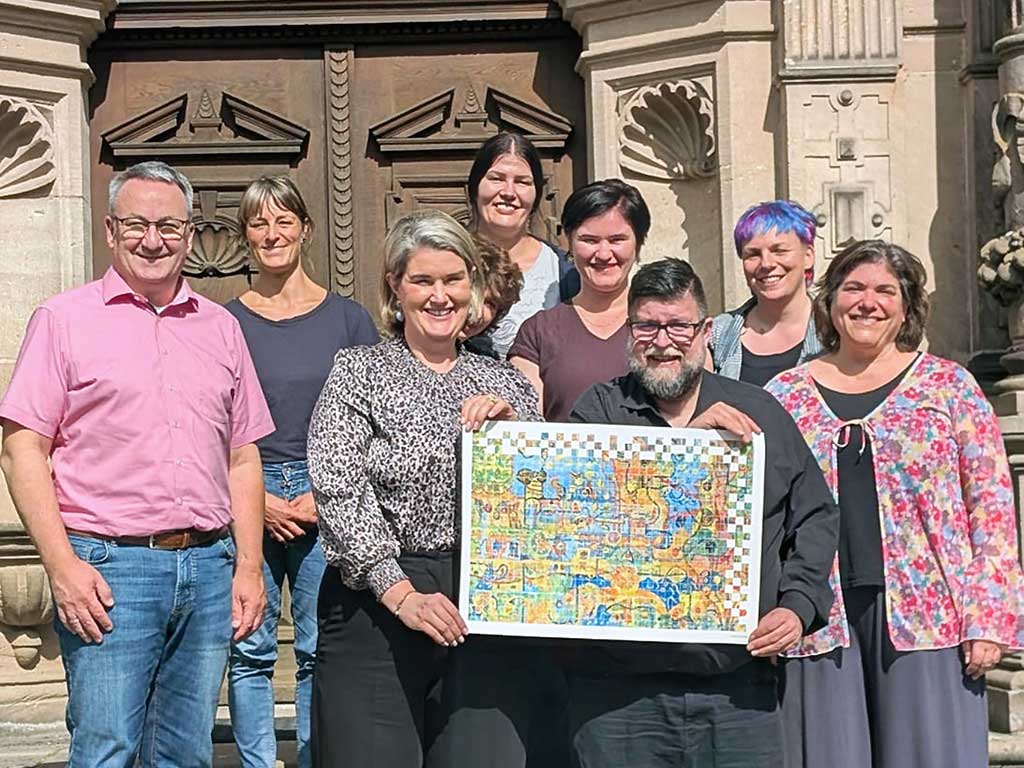 Auf einer Treppe steht eine Personengruppe in 2 Reihen. Die beiden Personen in der vorderen Reihe halten gemeinsam eine große bunte Urkunde in den Händen. Im Hintergrund sieht man eine schöne verzierte Eingangstür aus dunklem Holz eines alten Gebäudes.