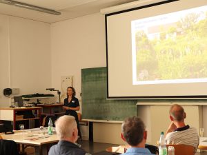 Die Referentin steht im Veranstaltungsraum vor der Leinwand, auf der ein Bild von ihrem Garten zu sehen ist.