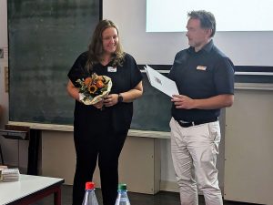 Die zu ehrende Person steht mit einem Blumenstrauß links neben dem Überbringer, der ihr gerade eine Ehrenurkunde überreicht.
