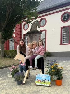 3 Mädchen sitzen auf einem Holzpferd, dahinter steht eine Frau und im Vordergrund sieht man einen Lernkoffer auf dem Pferde abgebildet sind. Rechts und links stehen verschiedene Blumentöpfe mit bunten Blumen.