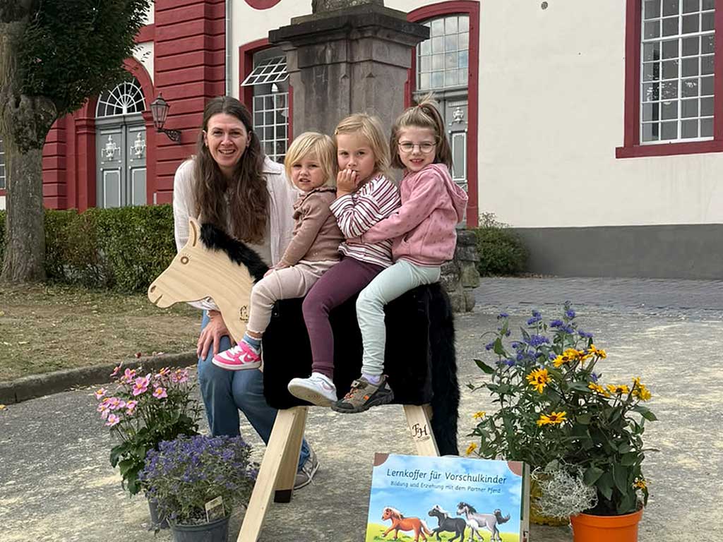 3 Mädchen sitzen auf einem Holzpferd, dahinter steht eine Frau und im Vordergrund sieht man einen Lernkoffer auf dem Pferde abgebildet sind. Rechts und links stehen verschiedene Blumentöpfe mit bunten Blumen.