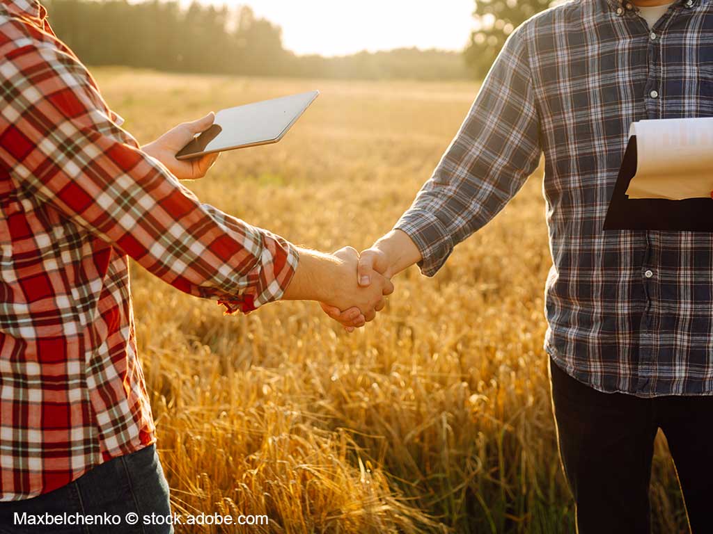 Detailfoto von 2 männlichen Personen in karierten Hemden, die sich die Hand reichen; im Hintergrund ein Getreidefeld im Sommer