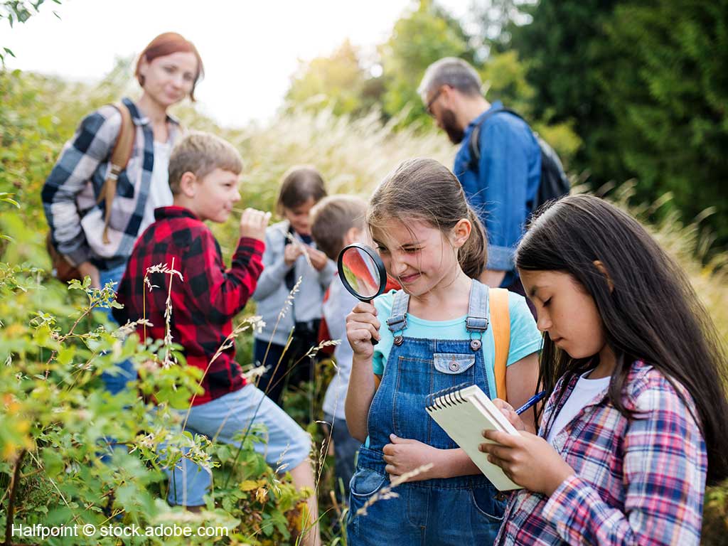 Personengruppe (5 Kinder und 2 Erwachsene) stehen auf einer Wiese; im Vordergrund sieht man 2 Mädchen; ein Mädchen schaut sich Gräser mit einer großen Lupe an; das andere Mädchen macht sich Notizen auf einem Schreibblock