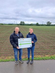 Landwirt Wolfang Wegfahrt und LeguNet Regionalmanagerin Clara Stieg stehen vor dem Sojabohnenfeld, auf dem sich die Sortendemo befindet. Die Sojabohnen sind noch klein. Sie halten gemeinsam ein Schild mit dem Logo des LeguNet und der Aufschrift: Soja-Sortendemonstration.